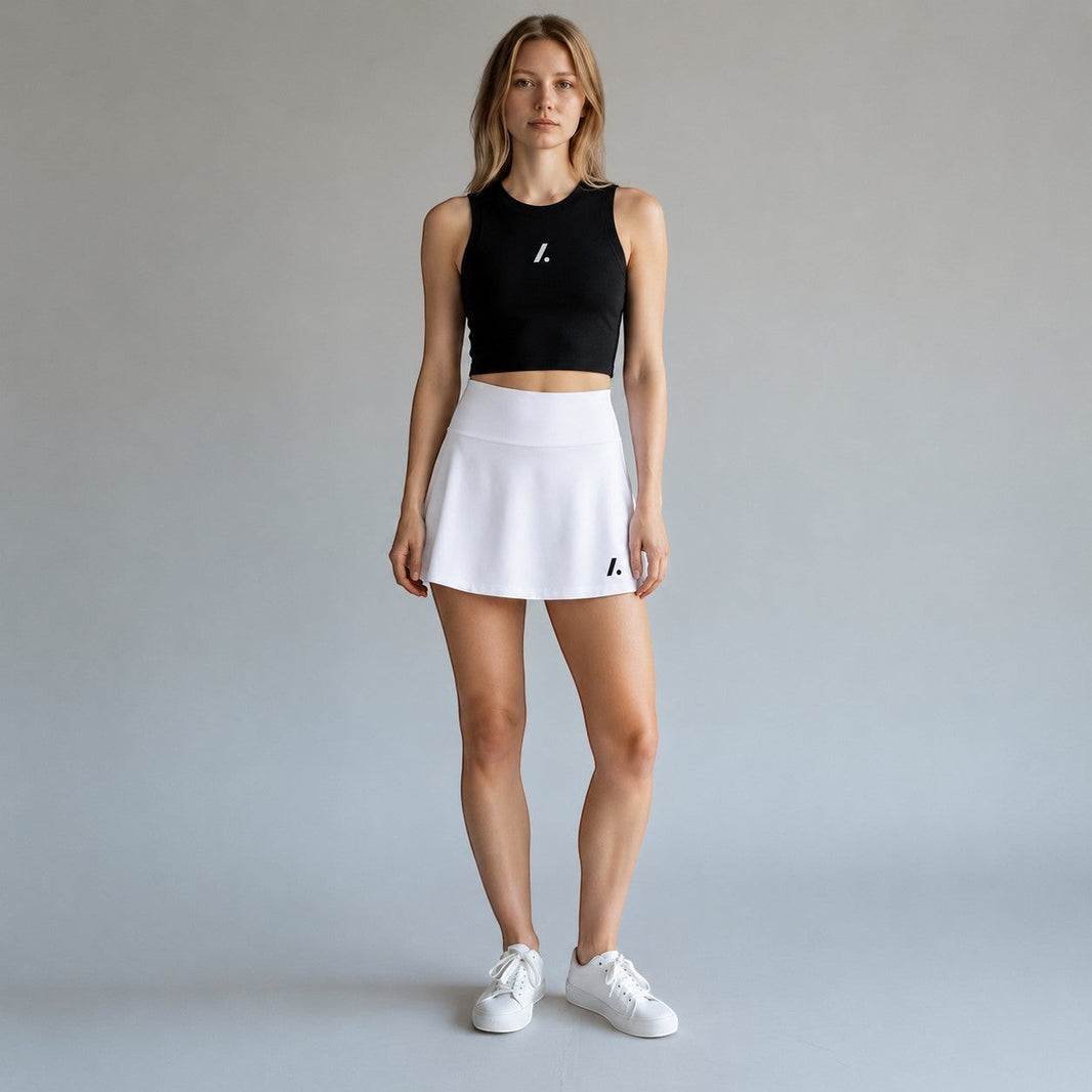 TwentyTenCollective white athletic skort with built-in shorts and flattering A-line skirt. Model wears black tank top and white sneakers.
